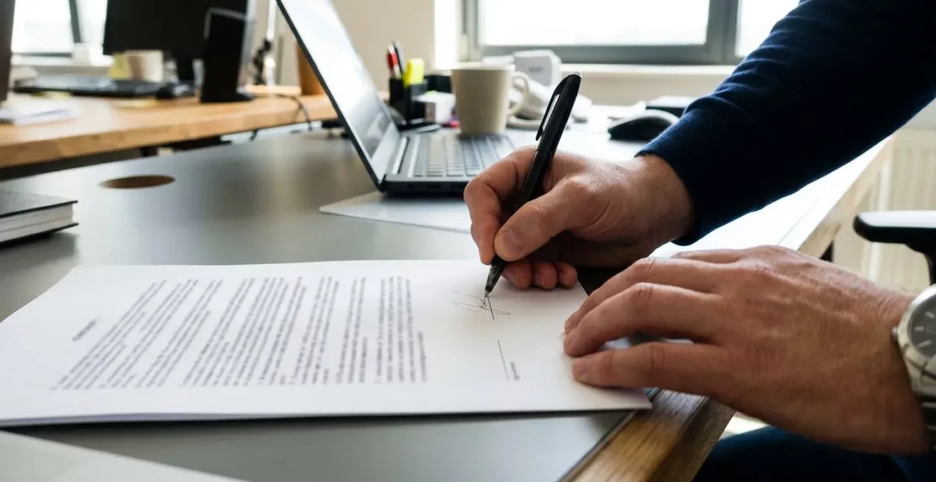 Gros plan sur des mains signant un document contractuel avec un stylo noir, le texte du contrat apparaît totalement flouté